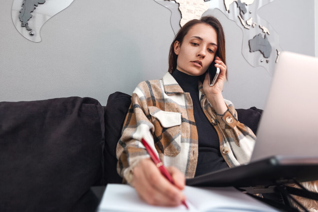 young woman working laptop writing note notebook talking cell phone